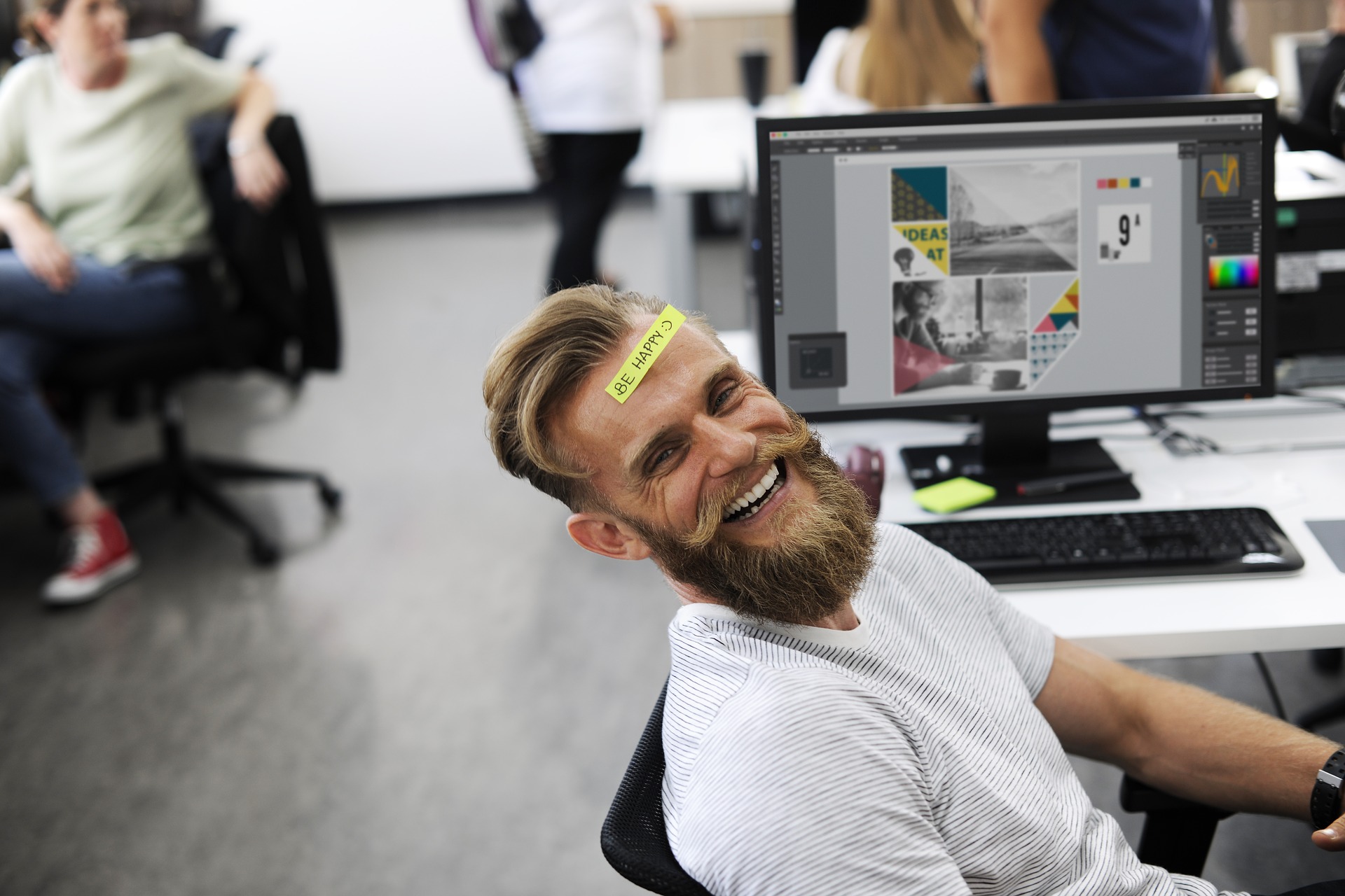 Bearded employee smiling at desk with "be happy" written on forehead