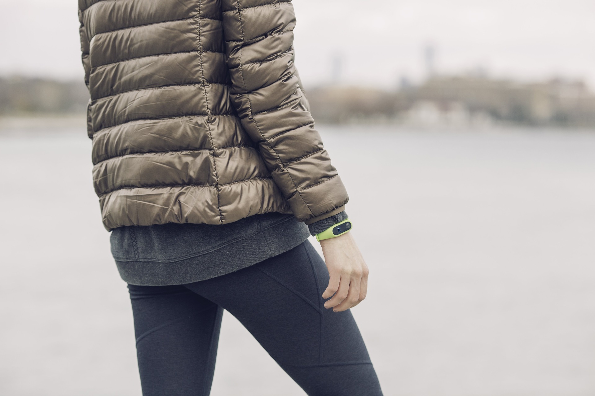 Woman in leggings and brown puffy coat standing next to water with fitness tracker on wrist