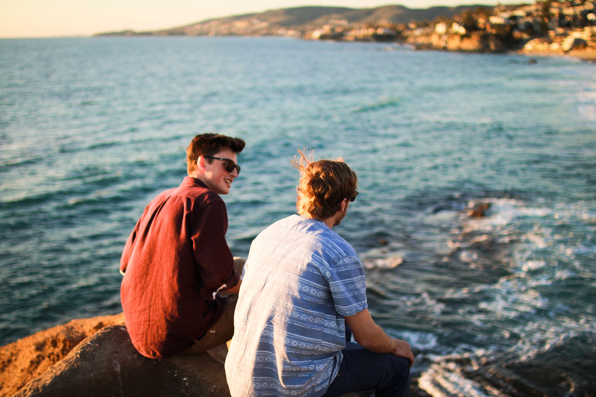 Two young men talking while sitting on rock beside body of water