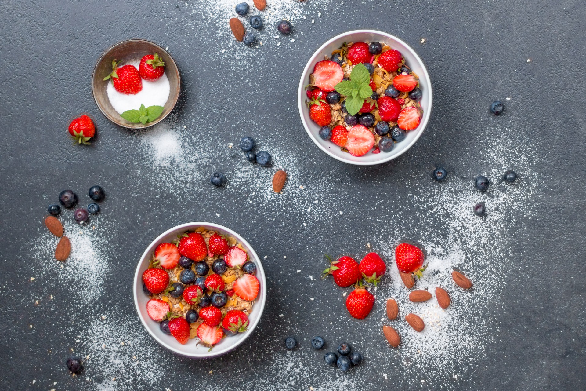 Overhead view of strawberries, raspberries, blueberries, and almonds in small bowls on blue table