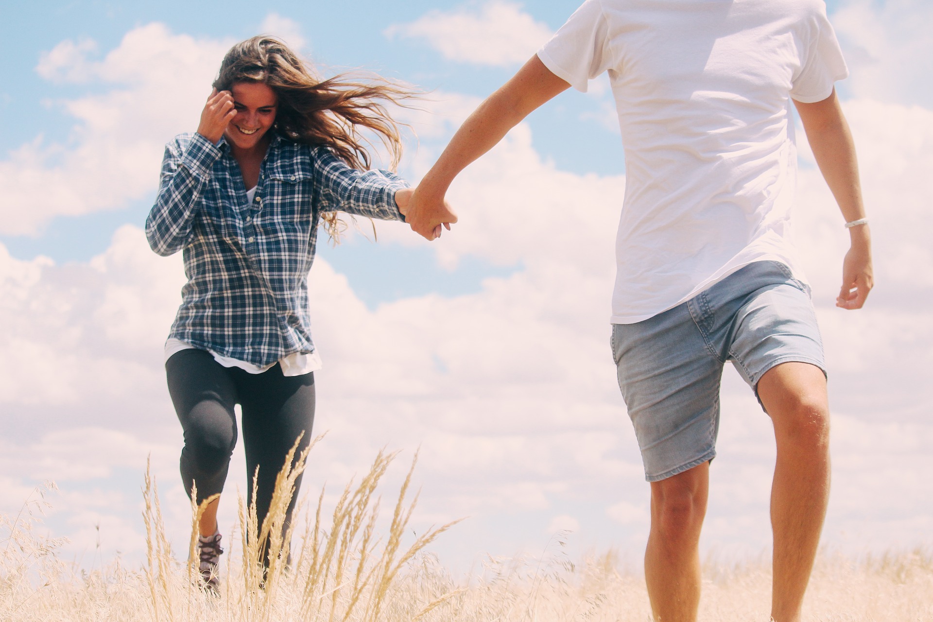 Man and woman holding hands while walking through field of tall grass with clouds in the blue sky