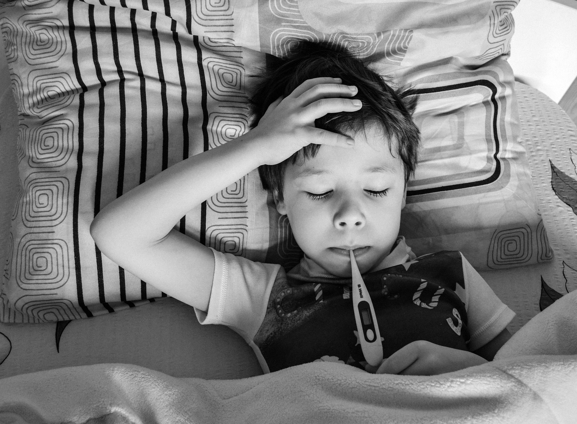 Black and white photo of a boy laying in bed with his hand on his forehead and a thermometer in his mouth